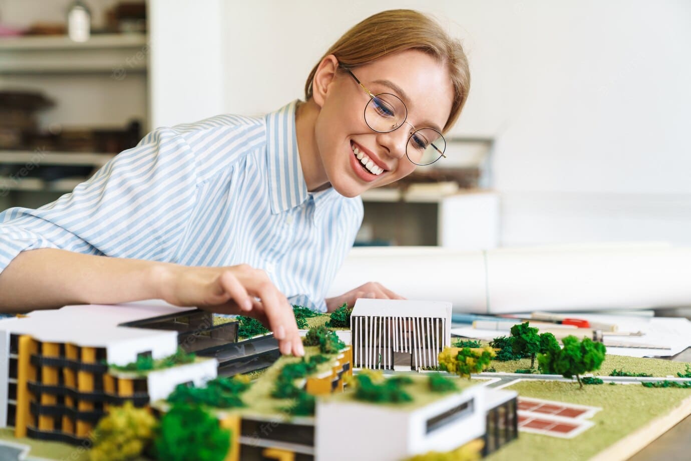 joyful-young-woman-architect-in-eyeglasses-designing-draft-with-house-model-and-sitting-at-workplace_171337-117656